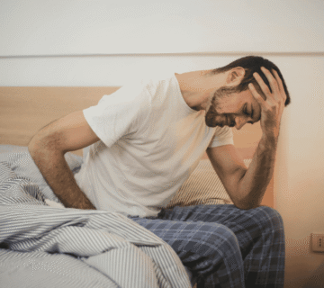 Pessoa sentada na cama em um quarto, com a mão na testa, demonstrando sinais de exaustão mental, fadiga e esgotamento emocional 