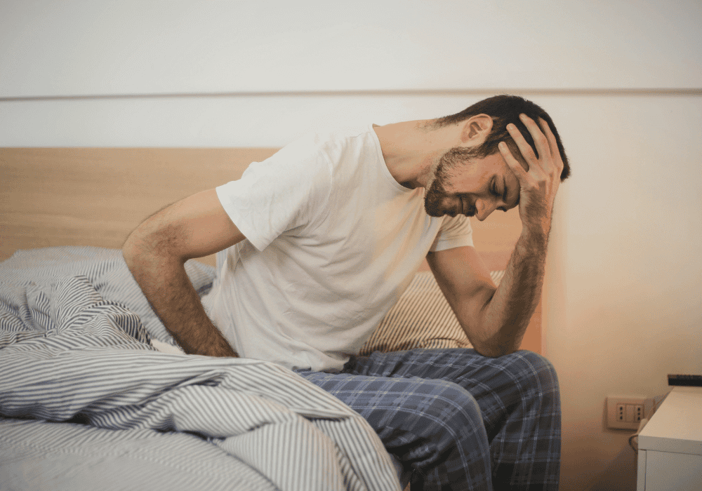 Pessoa sentada na cama em um quarto, com a mão na testa, demonstrando sinais de exaustão mental, fadiga e esgotamento emocional 